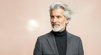 Mature man with gray hair and beard wearing charcoal blazer and black turtleneck, smiling against neutral beige background for corporate and lifestyle use.