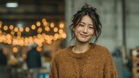 Young Asian woman smiling warmly in cozy brown sweater with messy bun hairstyle against blurred cafe lights background for lifestyle and portrait concepts.