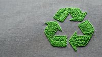 Green recycling symbol made from textured leaves on gray fabric background representing eco-friendly sustainability and environmental conservation concepts.