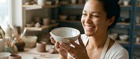 Young woman of East Asian descent, potter and ceramicist, smiling while holding handmade ceramic bowl in studio workshop with clay vessels and tools.