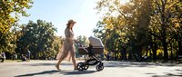 Woman with long hair wearing casual clothing pushing a baby stroller through a sunny park with golden autumn foliage, ideal for parenting and lifestyle content.
