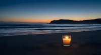 Glowing candle on sandy beach at twilight with ocean waves and mountain silhouette creating peaceful evening atmosphere for relaxation and meditation.