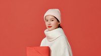 Young Asian woman in white knit hat and scarf holding coral gift box against matching red background, creating stylish monochromatic holiday portrait.
