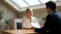 Mature woman with blonde hair and glasses, wearing beige blazer, conducting job interview with male candidate in modern office with skylight and potted plants.