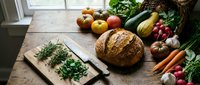 Fresh organic vegetables, fruits and artisan bread arranged on rustic wooden table near window. Healthy farm-to-table ingredients for cooking and meal preparation.