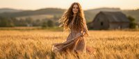 Young woman with long wavy hair in flowing dress sitting in golden wheat field at sunset with rustic barn and rolling hills in background.