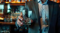 Professional businessman in elegant suit holding crystal whiskey glass in upscale bar setting with warm ambient lighting and blurred background.