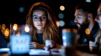Young Caucasian woman in glasses illuminated by laptop screen during late night meeting with colleagues, bokeh lights background creates atmospheric business setting.