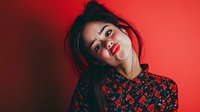 Young woman with messy updo hairstyle wearing floral pattern blouse against vibrant red background, showcasing confident expression.