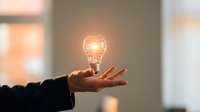 Silhouetted hand holding glowing incandescent bulb against blurred interior background, symbolizing innovation, creativity, and bright ideas for business concepts and educational materials.