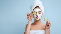 Young woman with facial mask holding cucumber slices for eye treatment, wearing white towel on head against blue background, puckering lips.