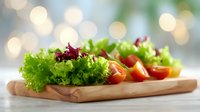 Fresh green lettuce leaves and red cherry tomatoes arranged on wooden cutting board against soft bokeh background, appetizing healthy salad ingredients.