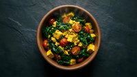 Vegetable stir-fry with corn, kale, and tomatoes in terracotta bowl on dark slate surface. Healthy meal preparation and nutritious cooking concept.