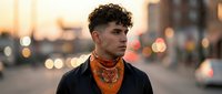 Young Hispanic man with curly hair wearing orange bandana and black jacket in urban street setting during golden hour sunset with bokeh lights.
