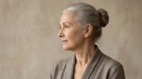 Elegant senior woman with gray hair in bun wearing beige blazer looking sideways in profile view against neutral background for business and lifestyle concepts.
