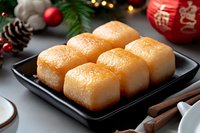 Fresh homemade sweet rice cakes on black ceramic plate with Christmas decorations, pine cones and cinnamon sticks against dark background with bokeh lights.