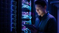 Young Asian male IT engineer smiling while using tablet in server room, managing network infrastructure with glowing rack equipment.