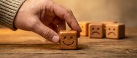 Hand selecting wooden cube with happy face from sad emotion blocks on table representing positive mindset choice and emotional wellbeing concept.