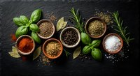 Aromatic herbs and spices collection with basil, rosemary, bay leaves, peppercorns, coriander seeds and sea salt arranged in wooden bowls on dark slate background.