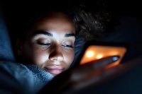Young mixed race woman illuminated by phone screen light while lying in bed at night, capturing modern digital lifestyle and screen time habits in darkness.