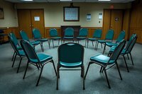 Empty chairs arranged in circle formation in meeting room with carpeted floor and wood paneled walls, suggesting group therapy, support group or team building space.