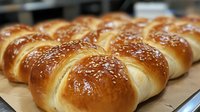 Fresh baked sesame bread rolls arranged in rows on parchment paper, showing golden brown crust and white sesame seeds sprinkled on top, bakery food photography.