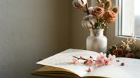Open journal with handwritten notes and delicate pink cherry blossoms, dried flowers in vase by window, soft natural light, cozy aesthetic.