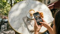 Person taking photo of pastry and coffee with smartphone on marble table outdoors, capturing food content for social media and blogging purposes.