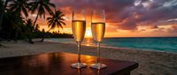 Two champagne flutes on a wooden table at a tropical beach sunset, with palm trees and vibrant sky, evoking romance and celebration.
