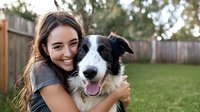 Young Hispanic woman with long dark hair embracing happy Border Collie dog outdoors in backyard garden, showing joyful genuine emotions and connection between pet and owner.