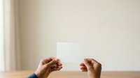 Two hands holding blank white card against neutral beige wall with natural light, suitable for mockups, presentations, advertising campaigns, and business communications.