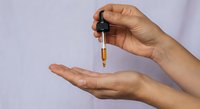 Woman hands applying essential oil serum from glass dropper bottle for skincare routine and natural beauty treatment on purple background.