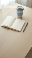 Open notebook with blank pages and pen on wooden desk next to reusable coffee cup, workspace setup for writing, planning, or creative work.