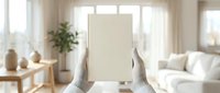 Hands holding blank cream colored card in bright modern living room with natural light, sofa, wooden furniture and potted plant for mockup and branding purposes.