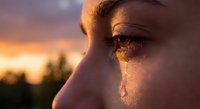 Close-up profile of young woman with tear rolling down cheek during golden hour sunset, emotional moment capturing sadness and contemplation outdoors.
