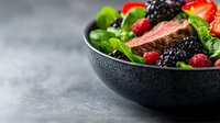 Fresh grilled beef steak slices with mixed berries, strawberries, blackberries and green salad leaves served in black ceramic bowl on gray background.