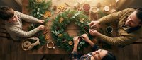Three people crafting a festive Christmas wreath with pine, dried oranges, and berries on a rustic wooden table, top view holiday scene.
