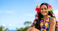 Young woman with long dark hair and medium skin tone wearing colorful flower lei garland around neck and in hair, smiling at camera outdoors with blue sky background.