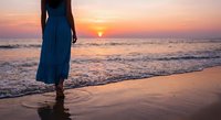Woman in blue dress walking on beach at sunset with golden sky and ocean waves creating peaceful vacation moment.