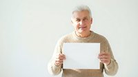 Senior man with white hair and light skin tone, wearing a beige ribbed sweater, holding blank white paper against white background. Suitable for business, advertising, and promotional campaigns.