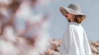 Woman in wide-brimmed beige hat and white flowing outfit standing against blurred natural background, creating elegant silhouette for fashion and lifestyle content.