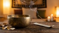 Tibetan singing bowl with burning incense stick creating smoke on wooden table in warm candlelit room for meditation and spiritual wellness practices.