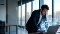 Focused Caucasian businessman in suit working on laptop with glowing digital holographic icons in modern office.