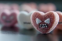 Pink sugar-coated heart candy with angry monster face expression and fangs against blurred background of similar sweets, macro photography for Valentine concept.