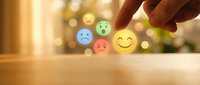Hand selecting happy yellow smiley face emoji from row of colorful emotion icons on wooden surface with warm bokeh background for feedback concept.