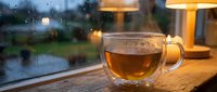 Steaming hot tea in transparent glass mug on wooden table by rainy window with warm lamp lighting creating cozy atmospheric indoor scene for relaxation.