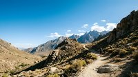 Scenic mountain landscape with winding dirt trail through arid valley, rocky peaks, and sparse vegetation under clear blue sky, suitable for travel and nature content.