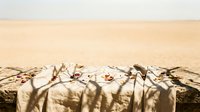 Elegant outdoor dining table setup on sandy beach with white linens, plates, and glassware for romantic seaside meal or coastal wedding reception.