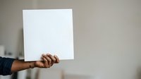 Adult male with medium brown skin tone holding blank white square sign or card against neutral beige wall background for messaging or advertising purposes.