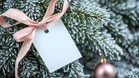 White gift tag with pink ribbon on snow-covered evergreen branches with rose gold ornament for holiday greeting cards and seasonal marketing materials.
